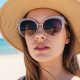 A woman wearing oversized cat-eye sunglasses on a sunny beach, styled with affordable luxury sunglasses and a wide-brim straw hat