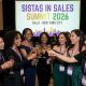 A group of women of color in professional attire networking and celebrating at the Sistas in Sales Summit 2026 Gala in New York City, with a vibrant event backdrop behind them